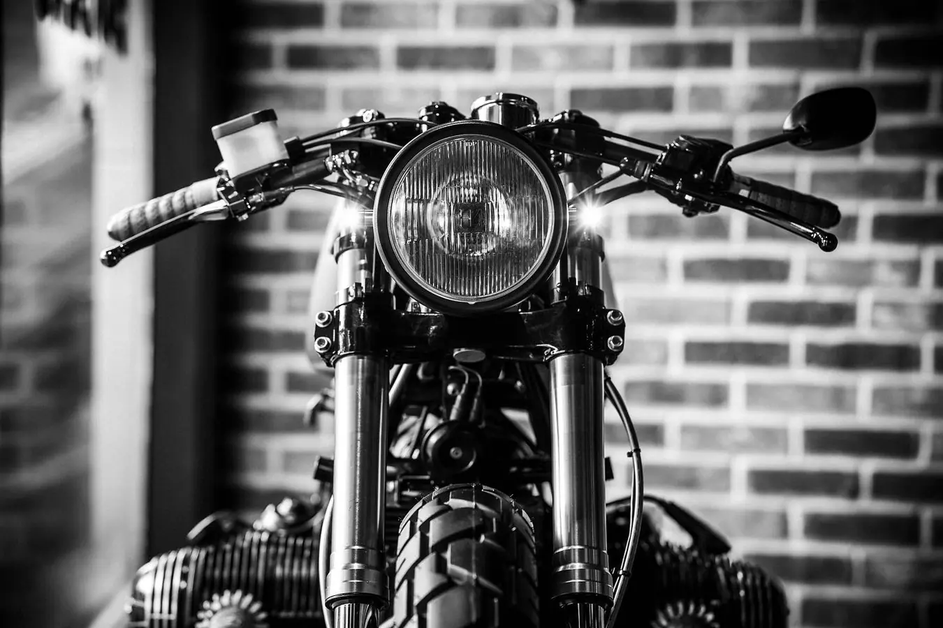 Black and white close-up of a boxer-twin cafe racer’s round headlight and bars against a brick wall